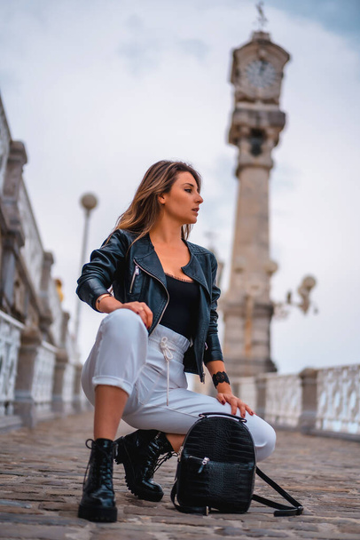 Lifestyle in the city with a blonde girl in white pants and a leather jacket. Crouched on a hill with city clocks in the background looking to the right - Foto, Imagem