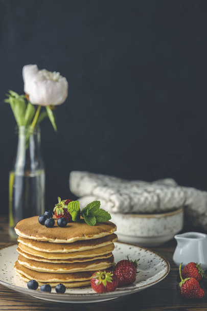 Pancake with strawberry, blueberry and mint in ceramic dish, syrup from small ceramic jar and flowers on a dark wooden table and black background.  - Foto, Imagen