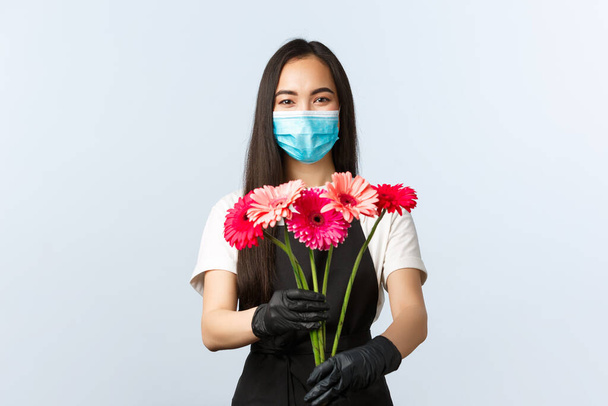 Small business, coronavirus pandemic, orders and shops concept. Smiling attractive female florist, store owner or employee in medical mask and protective gloves, giving customer purchased bouquet - Foto, immagini