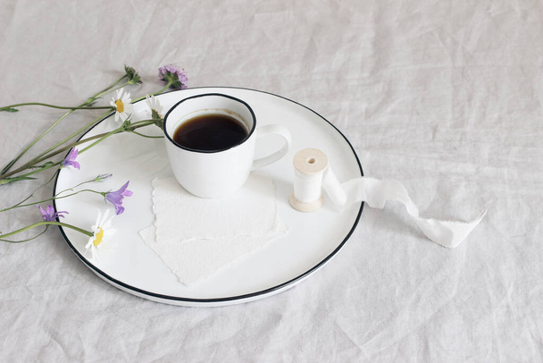 Spring, summer still life scene. Blank cotton paper greeting cards mockups on ceramic plate. Cup of coffee, silk ribbon, daisies and bluebells flowers on linen table cloth. Floral composition, top - 写真・画像