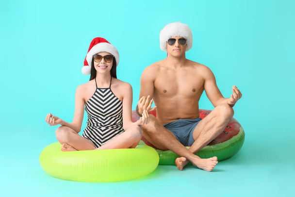 Meditating young couple with inflatable rings and Santa hats on color background - Photo, Image