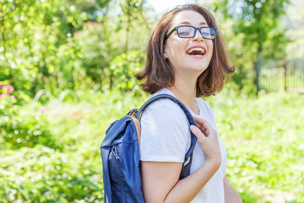 緑の公園の背景に笑みを浮かべてバックパック付き眼鏡で幸せな美しい正の学生の女の子。昼休みに学内で休んでいる女性。教育とレジャーの概念 - 写真・画像