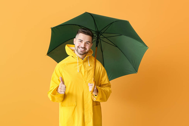 Young man in raincoat and with umbrella showing thumb-up on color background - Фото, изображение