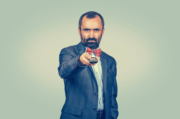 Adult displeased bearded man in elegant suit with red bow tie holding remote tv control. Mixed race bearded model isolated on green yellow uniform gradient background. Positive emotion - Fotoğraf, Görsel