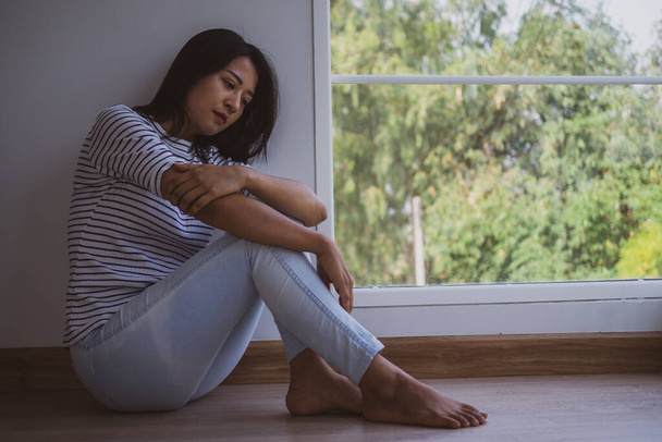 Una mujer asiática sintiéndose triste y sola sentada junto a la ventana de la casa. Confusión, decepción y problemas mentales - Foto, Imagen