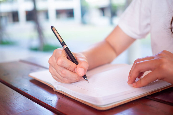 Cerca de las manos de una mujer sosteniendo un bolígrafo para escribir en un cuaderno sentado en una mesa fuera del lugar
. - Foto, imagen