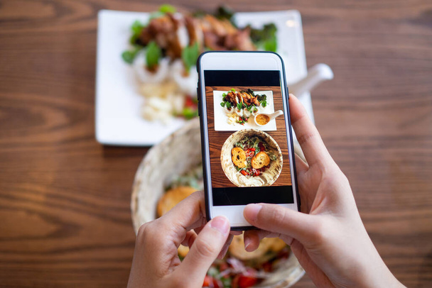 Ręka kobiety używającej telefonu komórkowego do robienia zdjęć jedzenia na stole, aby przeglądać i przesyłać je do portali społecznościowych. - Zdjęcie, obraz