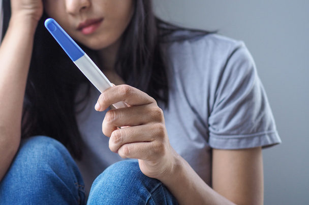 Asyalı kadın, hamilelik testini elinde tutarken üzgün ve korkmuş hisseder ve yatak odasında olumlu sonuç verir. Beklenmedik hamilelik ve kürtaj kavramı - Fotoğraf, Görsel