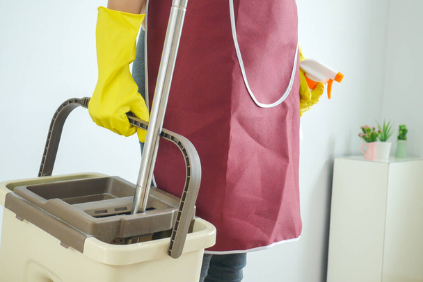 De huisvrouw draagt schoonmaakmiddelen, draagt rubberen handschoenen en hijst de emmer water. Reinigingsconcept - Foto, afbeelding