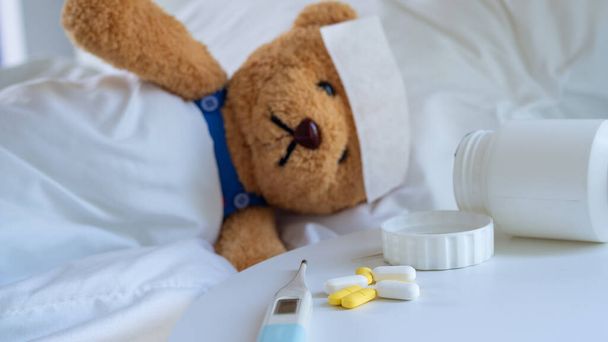 Teddy urso está doente na cama febre alta e tem um lençol de redução de febre na testa. Ao lado da cama estavam o termômetro e a medicação
. - Foto, Imagem