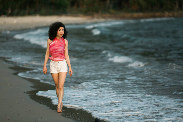 Mixed race woman walks along the edge of the surf on the sea beach. - Foto, afbeelding