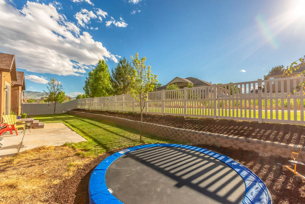 Trampoline op zonovergoten achtertuin van thuis met patio en plantbed tegen hek - Foto, afbeelding
