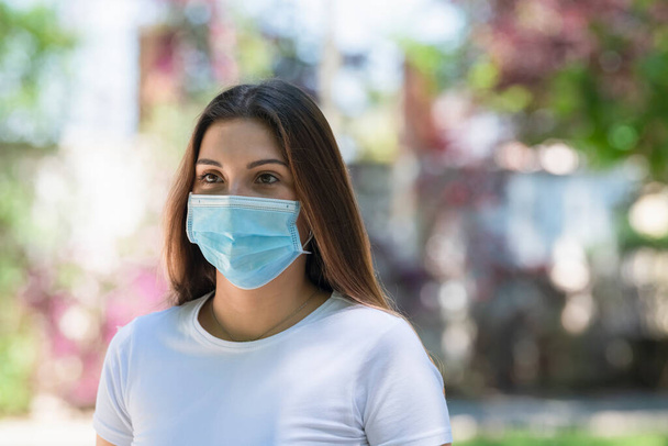 Close up of a young beautiful woman wearing a surgical mask outdoors on an out of focus background. New lifestyle and safety concept. - Foto, Imagem