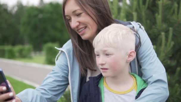 Eine Mutter und ihr Teenager-Sohn machen ein Selfie mit ihrem Handy, während sie in einem Park spazieren gehen - Filmmaterial, Video