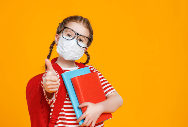 Розумна дівчина в масці і окулярах обіймає підручники і показує тумблери під час навчання в школі під час пандемії проти жовтого фону
 - Фото, зображення