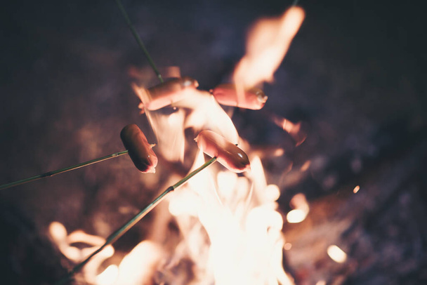 キャンプファイヤーの上でホットドッグソーセージを作って調理する。木の枝にたき火の炎の上に食べ物を焼く-自然の中でスティック槍a - 写真・画像