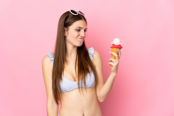 Young caucasian woman in swimsuit holding a cornet ice cream isolated on pink background with sad expression - Фото, зображення