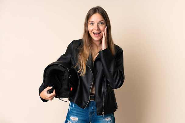 Young Lithuanian woman holding a motorcycle helmet isolated on beige background with surprise and shocked facial expression - 写真・画像