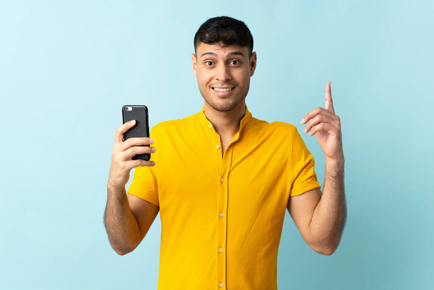 Jonge Colombiaanse man met behulp van mobiele telefoon geïsoleerd om blauwe achtergrond wijzen op een geweldig idee - Foto, afbeelding