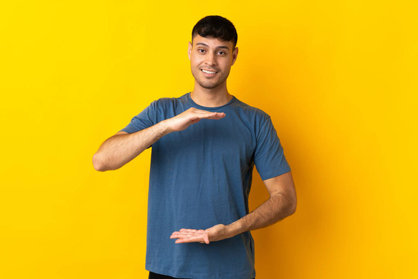 Jovem colombiano isolado em fundo amarelo segurando espaço imaginário na palma da mão para inserir um anúncio
 - Foto, Imagem