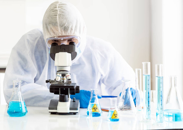 Young Asian scientist doing some research and  looking through a microscope in a laboratory to study about coronavirus COVID-19 to develope vaccine for 2019-ncov world pandemic. Healthcare concept. - Photo, image