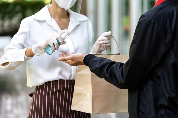 Waitress spraying alcohol on hand of delivery man before give bakery grocery bag for cleaning and hygiene as new normal for Food delivery service while coronavirus covid-19 pandemic. - Foto, afbeelding