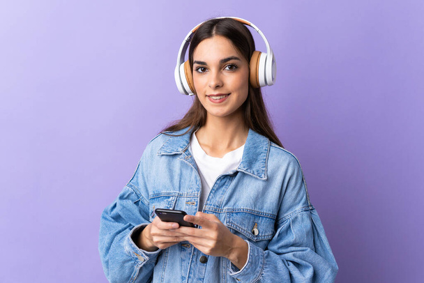 Jovem caucasiana isolada no fundo roxo ouvindo música com um celular e olhando para a frente
 - Foto, Imagem