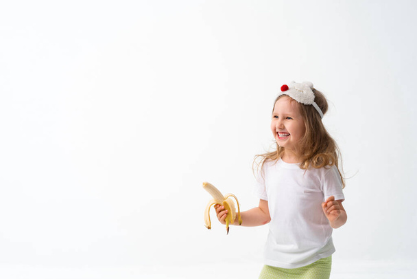 Cute little 4 year old Caucasian girl in casual clothes, holding a banana in her hand, looks very happy, isolated on a white background. concept of natural nutrition, the benefits of fruit. Copy spase - Photo, Image