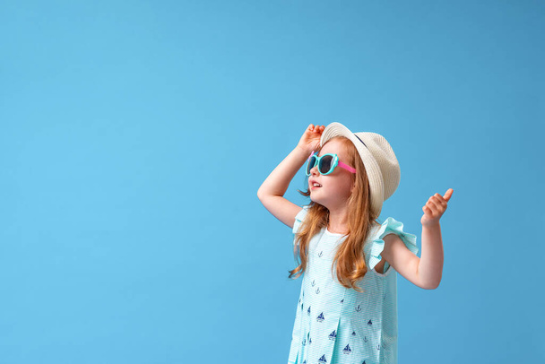 a cute little girl of 4 years old, in a dress, hat and sunglasses, poses looking away, on a blue background. The child will be happy to start the summer holidays and go on a trip - Foto, Imagen