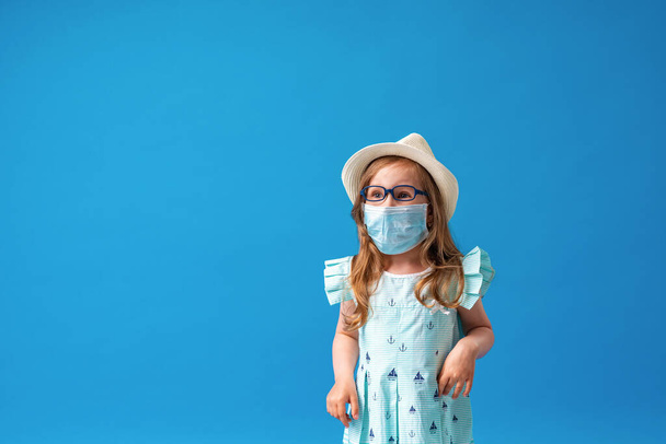 small cheerful 4-year-old girl in glasses, hat, and summer dress poses on blue background. child with a face protection mask due to air pollution or a coronavirus epidemic. Rest during pandemic. - Foto, Bild