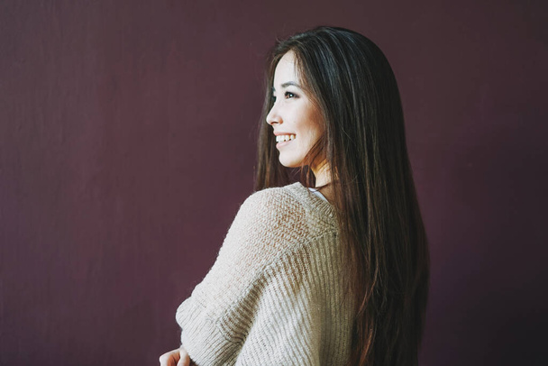Charming smiling asian girl with long brown hair in beige sweater isolated on the dark bordo background - Photo, Image