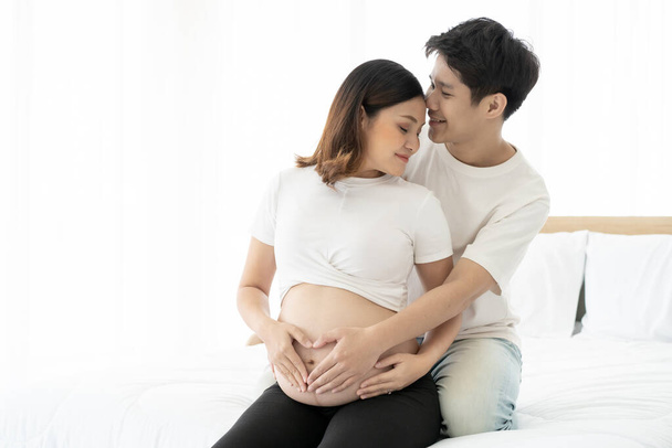 Asian lovely husband and wife portrait, his wife is in pregnancy. Husband tenderly hug and kiss his wife on the bed with care. Husband embracing his pregnant wife and making their hands in heart shape - Fotoğraf, Görsel