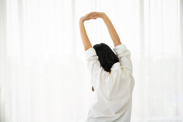 Hezká asijská žena v bílé košili, aby se protáhnout po probuzení z postele v dopoledních hodinách, volný čas a pohodlný životní koncept. - Fotografie, Obrázek