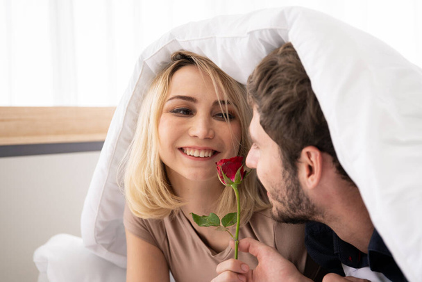 Young Western couple having a romantic moment together in bedroom, man giving a beautiful red rose to his wife in Valentine's day. - Φωτογραφία, εικόνα