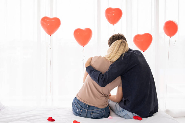 Ρομαντική στιγμή του νεαρού δυτικού ζευγαριού, ένα ζευγάρι κάθεται στο κρεβάτι στην κρεβατοκάμαρα με μπαλόνια κόκκινο σχήμα καρδιάς στο παρασκήνιο. Φωτογραφία με χώρο αντιγραφής. - Φωτογραφία, εικόνα