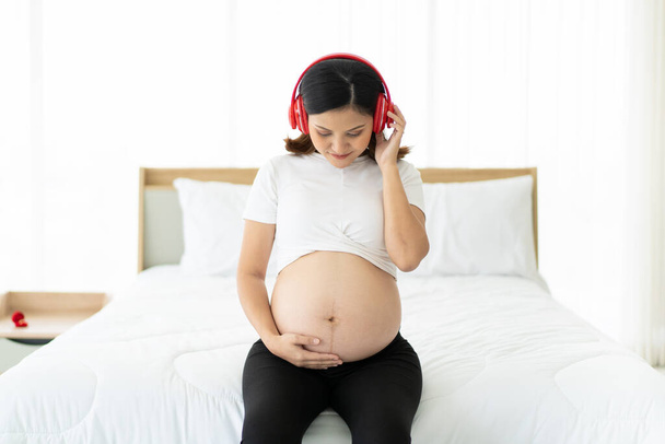 Happy pregnant woman relaxing in her bedroom and listening a music by using wireless headphones. Big stomach of pregnant woman sitting on the bed while listening a beautiful song. Wellbeing concept. - Φωτογραφία, εικόνα