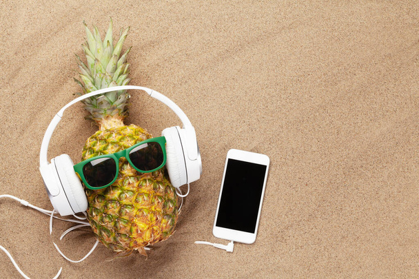 Ananas mûr avec lunettes de soleil et écouteurs sur fond de sable avec smartphone. Concept de voyage et vacances. Vue du dessus plat avec espace de copie
 - Photo, image