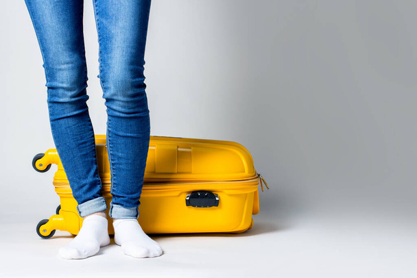 Naispuoliset jalat farkuissa lähellä keltaista matkalaukkua kevyellä taustalla. - Valokuva, kuva