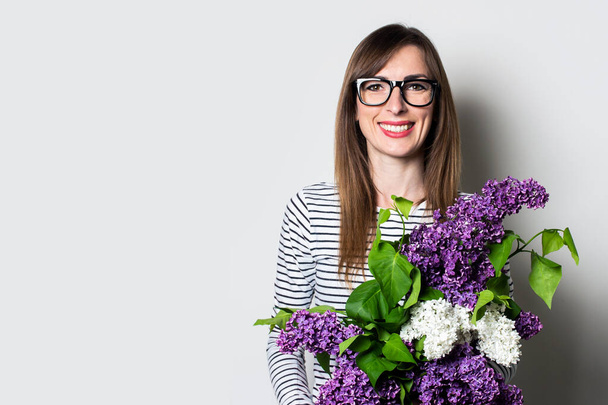 Friendly young woman in glasses holds a bouquet of lilacs on a light background - Photo, Image