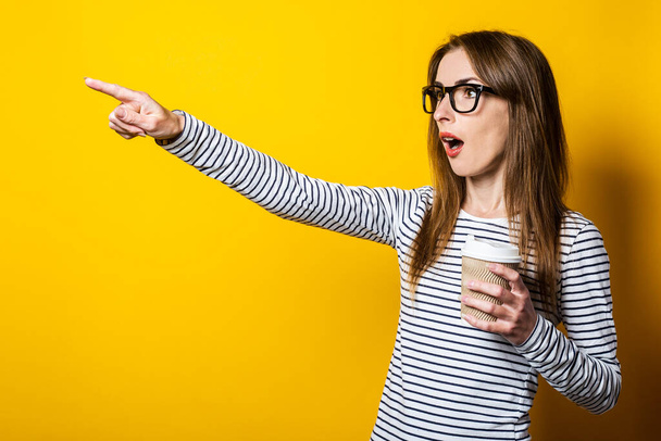 Młoda kobieta zaskoczona szklankami z kawą wskazuje palcem na bok na żółtym tle. - Zdjęcie, obraz