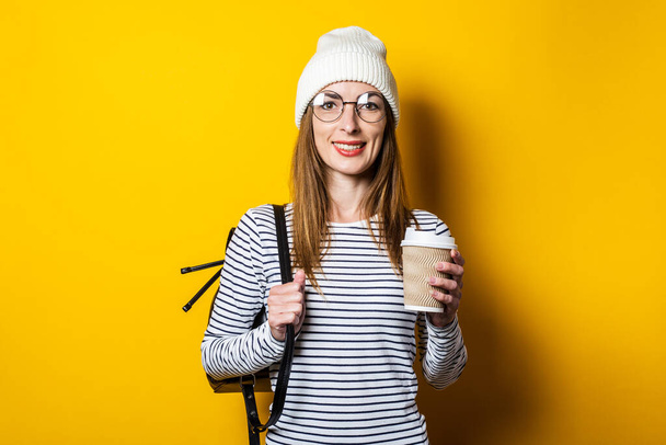 Портрет дружелюбной девушки в очках и шляпе, держащей бумажную чашку с кофе на желтом фоне
. - Фото, изображение