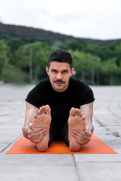 Jeune latin gay faire de l'exercice de yoga dans un parc. Tenir les jambes avec les mains et regarder la caméra. Pose de yoga coudée vers l'avant assise ou Paschimottanasana - Photo, image