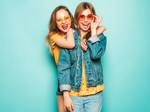 Two young beautiful smiling hipster girls in trendy summer yellow T-shirts and jeans jacket. Sexy carefree women posing near blue wall. Trendy and positive models having fun - Foto, Imagen