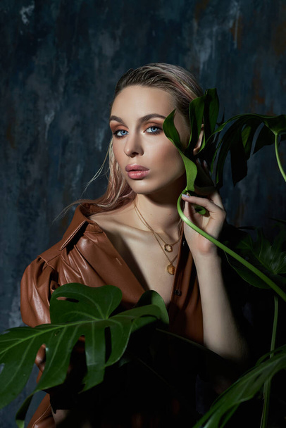 Portrait of a woman in tropical foliage. Wet hair, perfect figure and makeup. Beautiful blonde girl in leather dress and monstera leaves - Fotó, kép