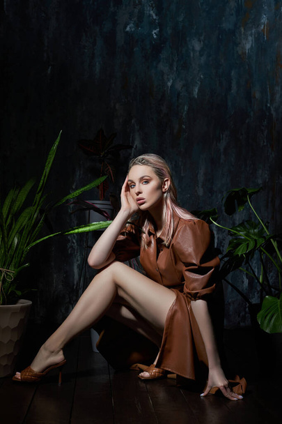 Portrait of a woman in tropical foliage. Wet hair, perfect figure and makeup. Beautiful blonde girl in leather dress and monstera leaves - Foto, afbeelding
