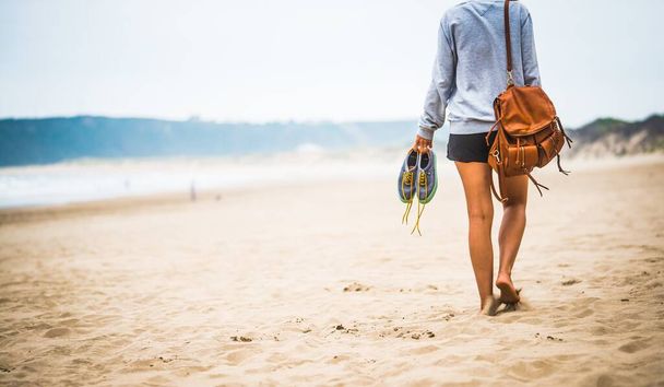 A young female with a brown backpack holding her shoes and walking through the beach under the sunlight - Foto, Imagem
