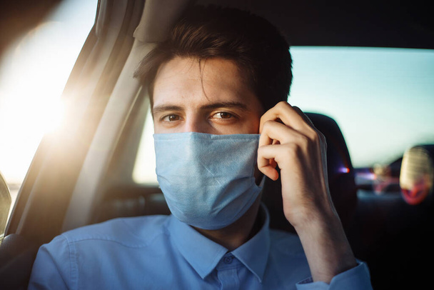 Młody biznesmen bierze taksówkę i patrzy przez okno samochodu na sterylną maskę medyczną. Mężczyzna siedzi na tylnym siedzeniu taksówki i przejeżdża się podczas pandemii koronawirusowej. Pojęcie odległości społecznej - Zdjęcie, obraz