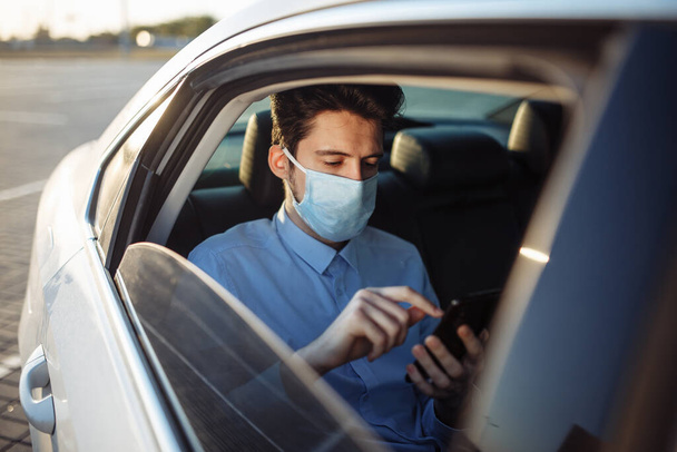 Young businessman takes a taxi and looks into his cell phone wearing sterile medical mask. A man sits on the back seat of taxi and takes a ride during coronavirus pandemic. Social distance concept - Photo, Image