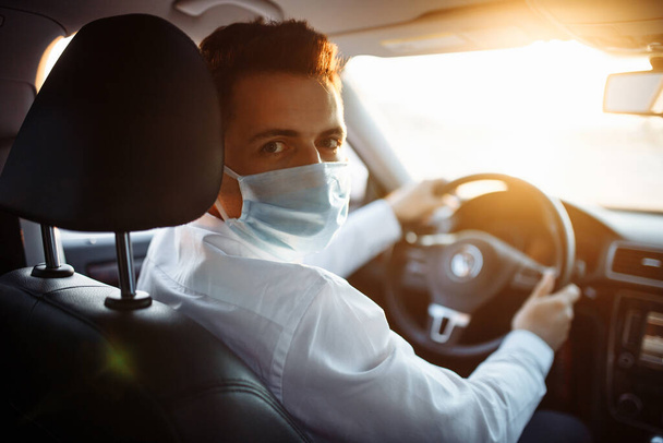Young man taxi driver steers the car during coronavirus pandemic wearing sterile medical mask. A boy drives a car through the traffic on the sunset. Social distance and health care concept - Photo, Image