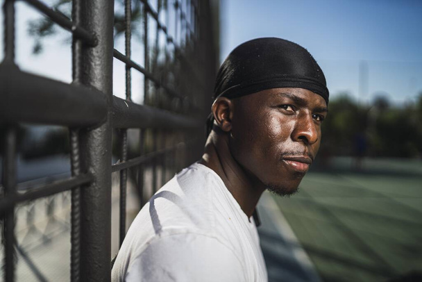 A closeup shot of an African-American male in a white shirt leaning on a fence at the basketball court - Fotoğraf, Görsel